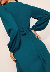 Mėlynas tekstūruotas suknelė su laisvu siluetu, turinti ilgas pūstas rankoves ir surištą juosmens detalę, išryškinanti glotnią ir šiek tiek susiraukšlėjusią medžiagą.