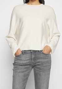 Suéter de color crema, de gran tamaño, con cuello redondo y mangas largas, combinado con jeans de mezclilla gris de corte clásico y con bolsillos delanteros.
