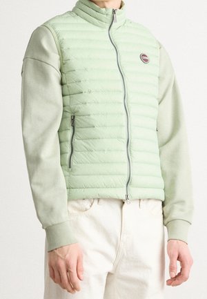 Personne portant un gilet matelassé vert clair avec fermeture éclair et logo sur la poitrine, par-dessus une chemise à manches longues assortie et un pantalon blanc cassé.