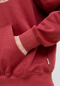 Sweatshirt rouge avec une texture douce, doté d'une poche kangourou et de poignets côtelés. Comprend une petite étiquette d'accent sur le côté.