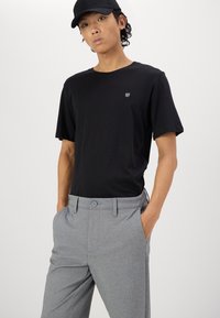 Schwarzes T-Shirt mit kurzen Ärmeln und kleinem Logo auf der Brust, kombiniert mit hellgrauen strukturierten Hosen mit Knopfverschluss und Seitentaschen.
