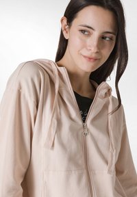 Sudadera con cremallera de color rosa claro con una textura suave, capucha ajustable y bolsillos canguro. La cremallera cuenta con un tirador circular plateado.