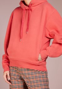 Koraalrode trui met capuchon, voorzien van trekkoorden, zijzakken en geribbelde manchetten. Gecombineerd met geruite broek.