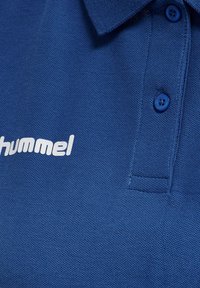 Blå polo skjorta med en texturerad tyg, med krage, två blå knappar och en vit logotyp på framsidan.