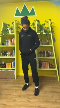 Zwarte hoodie met logo, bijpassende zwarte joggingbroek en zwarte schoenen. Houten boekenkast met boeken en planten in een gele kamer. Groene bergmuur.