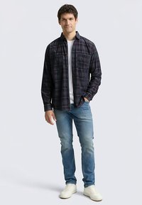Camicia di flanella a quadri scura con tonalità di blu, rosso e verde, due tasche sul petto; abbinata a jeans azzurri e sneakers bianche.