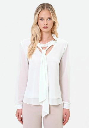 Donna bionda che indossa una camicetta bianca a maniche lunghe con dettaglio di sciarpa annodata sul collo e pantaloni beige chiaro.