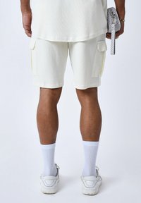 Pantalones cortos de cargador blancos con una tela texturada, bolsillos laterales y un ajuste relajado, combinados con calcetines blancos y zapatillas. Vistos desde atrás.