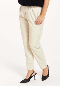 Beige faux leather pants with a high waist, pleated front, tapered legs, and side pockets, paired with black slingback heels.