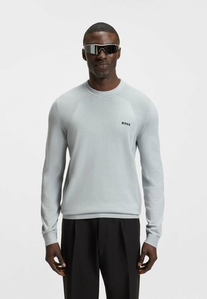 Hellgrauer Strickpullover mit gerippten Details an den Schultern, einem Rundhalsausschnitt und dem BOSS-Logo in Dunkelblau. Kombiniert mit schwarzen Hosen.