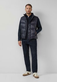 Gepolsterte marineblaue Weste mit hohem Kragen, Reißverschluss und vier vorderen Taschen, getragen über einem dunklen Hemd und Jeans, kombiniert mit hellen Sneakers.