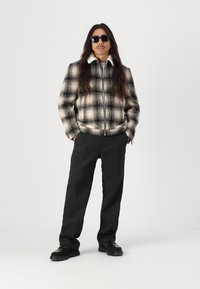 Kreminės ir juodos spalvos languota zip-up striukė; plati apykaklė ir ilgos rankovės. Pilkos kasdienės kelnės su šoninėmis kišenėmis. Juodi batai.
