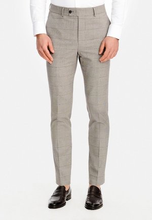 Mężczyzna ubrany w dopasowane, szare, w kratę spodnie wizytowe i czarne skórzane mokasyny, stojący z rękami swobodnie opuszczonymi wzdłuż boków.