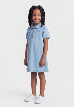 Uśmiechnięta młoda dziewczyna z warkoczami, ubrana w jasnoniebieską sukienkę z krótkim rękawem i białe trampki, stojąca na jednolitym tle.