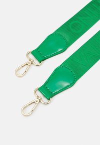 Grön textilrem med glänsande läderdetaljer och hårdvara i guldton. Graverad text på remmen ger textur och detalj.