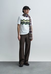 Λευκό πουκάμισο με κοντά μανίκια και ζιγκ-ζαγκ πράσινο και μαύρο μοτίβο, συνδυασμένο με καφέ παντελόνι φαρδιάς γραμμής και πολύχρωμο ριγέ κασκόλ.