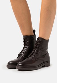 Bottes courtes en cuir marron avec fermeture à lacets sur le devant, fermeture éclair latérale, bout arrondi et petit talon bloc. Texture lisse avec détails minimalistes.