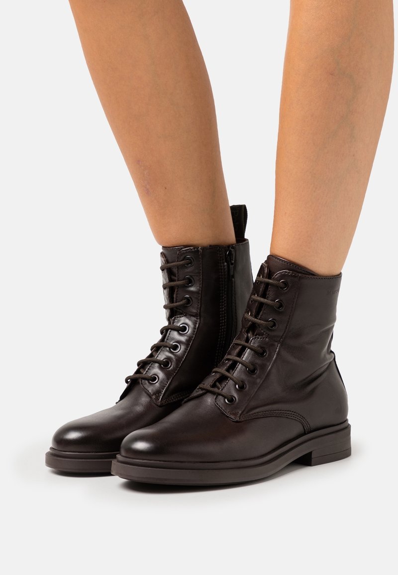 Bottes courtes en cuir marron avec fermeture à lacets sur le devant, fermeture éclair latérale, bout arrondi et petit talon bloc. Texture lisse avec détails minimalistes.