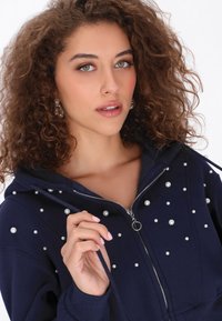 Granatowa bluza z kapturem na zamek z perełkowymi zdobieniami, regulowanym kapturem ze sznurkiem i miękką teksturą. Posiada przedni zamek błyskawiczny i luźny krój.