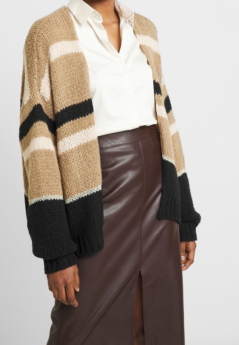 Femme portant un cardigan en tricot beige, noir et crème sur une chemise blanche, associé à une jupe taille haute en cuir marron foncé avec une fente à l'avant.