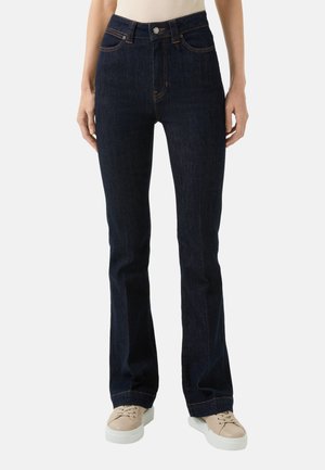 Jean en denim bleu foncé évasé avec une taille mi-haute, des coutures apparentes, cinq poches et une texture lisse, associés à des baskets beiges.