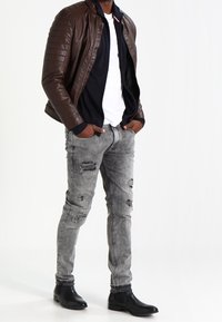 Hnědá kožená bomber bunda s prošívanými detaily, nošena přes bílé tričko. Šedé poškozené džíny a černé kotníkové boty dokončují vzhled.