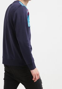 Uomo che indossa una felpa navy con dettagli floreali blu sulle spalle e jeans neri strappati, in piedi di lato contro uno sfondo bianco.