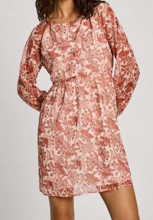 Vrouw draagt een jurk met lange mouwen en een lengte tot de knieën, met rode bloemenpatronen op een lichtroze achtergrond, staand met handen in haar zij.