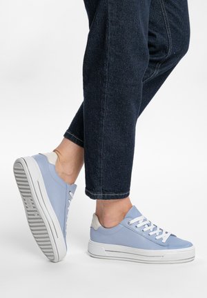 Osoba nosící světle modré tenisky s bílými podrážkami a tkaničkami, oblečená do tmavých ohrnutých džínů, stojící na jedné noze.