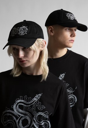 Kaks modelli, kes kannavad musti nokamütse ja särgikesi valge madu kujundusega, poseerivad tavalise halli tausta ees.