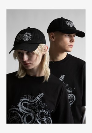 Twee modellen dragen zwarte petten en shirts met witte slangmotieven, poserend tegen een effen grijze achtergrond.