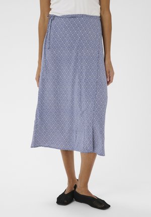 Femme portant une jupe midi à motifs bleus avec un lien sur le côté et des chaussures noires en tricot sans lacets, debout devant un fond uni.