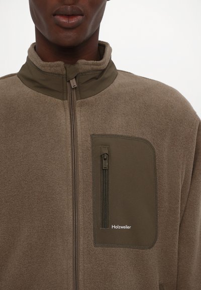 Fleece-Zip-Jacke in Beige und Olivgrün, mit hohem Kragen und einer mit Reißverschluss versehenen Brusttasche mit Logo. Weicher Stoff überall.