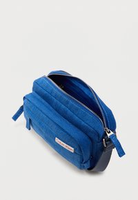 Μπλε τσάντα χιαστί από ύφασμα με φερμουάρ, υφή στην επιφάνεια και ετικέτα με λογότυπο. Διαθέτει κομψό ορθογώνιο σχήμα και πλαϊνές λαβές.