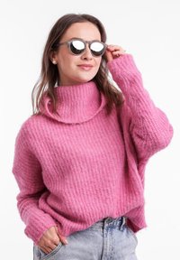 Růžový pletený svetr s vysokým rolákem a volným střihem. Modelka nosí kulaté šedé sluneční brýle, které ukazují texturovaný design látky.