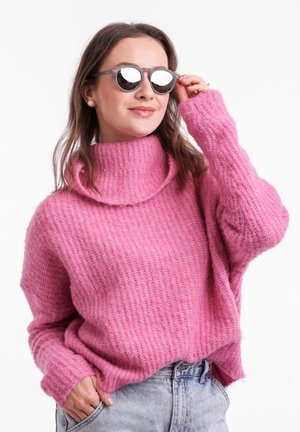 Růžový pletený svetr s vysokým rolákem a volným střihem. Modelka nosí kulaté šedé sluneční brýle, které ukazují texturovaný design látky.