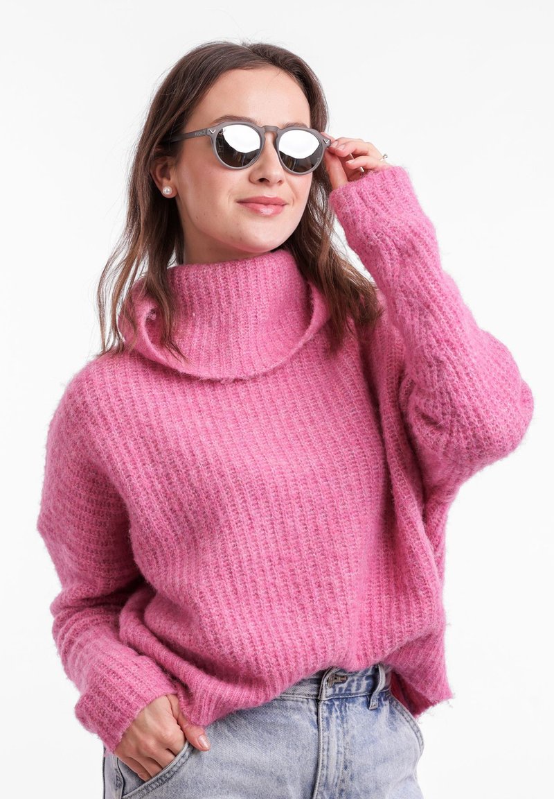 Růžový pletený svetr s vysokým rolákem a volným střihem. Modelka nosí kulaté šedé sluneční brýle, které ukazují texturovaný design látky.
