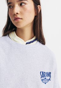 Harmaa huppari, jossa on sininen "SARDINE" -painatus ja kalagrafiikka vasemmassa rinnassa. Valmistettu pehmeästä materiaalista ja klassisessa crew neck -tyylissä.