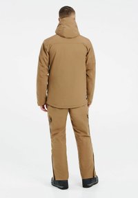 Chaqueta aislante marrón con capucha y pantalones a juego, que cuentan con bolsillos con cremallera y cremalleras laterales para ventilación. Textura mate.