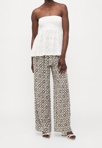 Haut sans bretelles blanc avec un motif brodé en œillets, associé à un pantalon large marron avec un motif géométrique en losanges. Sandales blanches.