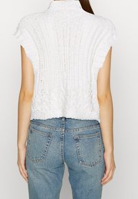 Bílý pletený crop top s žebrovanou texturou a vroubkovanými okraji, s vysokým límcem a krátkými rukávy. Kombinován se světle modrými džíny.