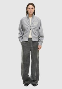 Grijze zip-up sweatvest met geribbelde manchetten, logo in lichtbeige, gecombineerd met wijde donkergrijze broek en zwarte schoenen.