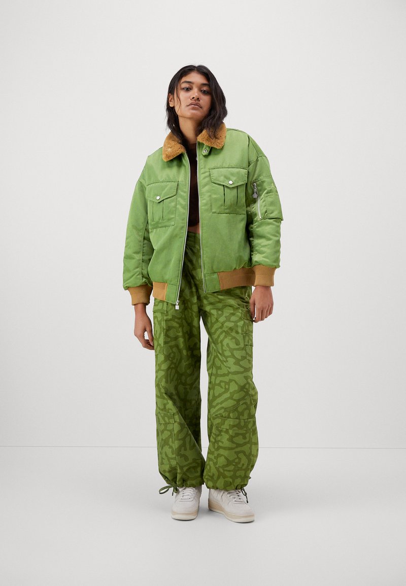 Donna che indossa una giacca bomber verde con colletto marrone e pantaloni cargo verde mimetico, in piedi davanti a uno sfondo chiaro e uniforme.