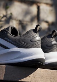 Paire de chaussures de sport grises et blanches étiquetées « Supernova Ease 2 » posées sur une surface en bois avec un arrière-plan extérieur flou.