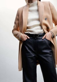 Abrigo beige a medida sobre un suéter de punto color crema, combinado con pantalones de cuero negro de cintura alta. Presenta doble botonadura y cinturón.