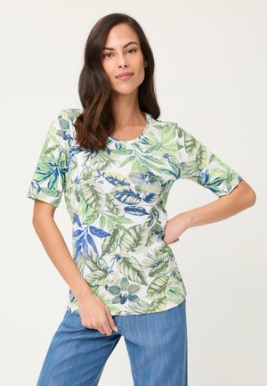 Kobieta z długimi ciemnymi włosami ubrana w białą koszulę z krótkim rękawem z niebieskim i zielonym motywem liściastego kwiatu oraz niebieskie dżinsy, pozująca na białym tle.