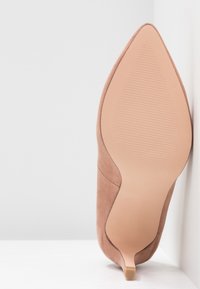 Chaussure à talon haut en daim beige à bout pointu, vue de la semelle avec un petit talon texturé sur fond blanc d’angle.