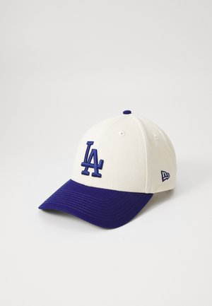Gorra de béisbol con una corona color crema y un visor púrpura, con un logotipo bordado "LA" en azul. Textura de tela suave y diseño estructurado.
