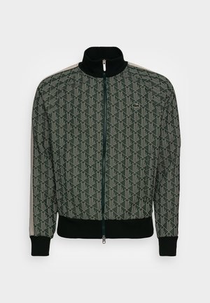 Veste zippée vert foncé avec motif géométrique, col et poignets noirs, rayures beiges sur les manches, et petit logo crocodile sur la poitrine.