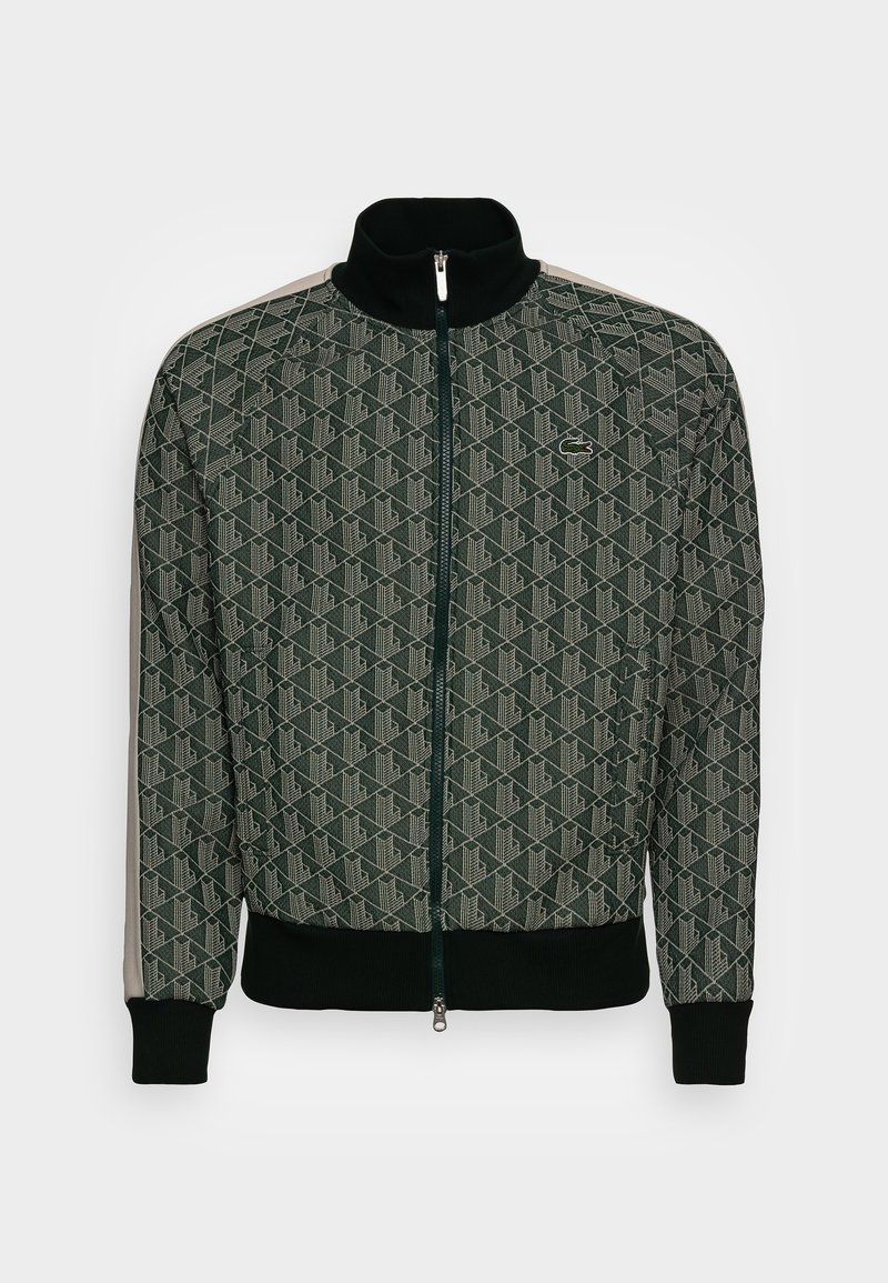 Veste zippée vert foncé avec motif géométrique, col et poignets noirs, rayures beiges sur les manches, et petit logo crocodile sur la poitrine.